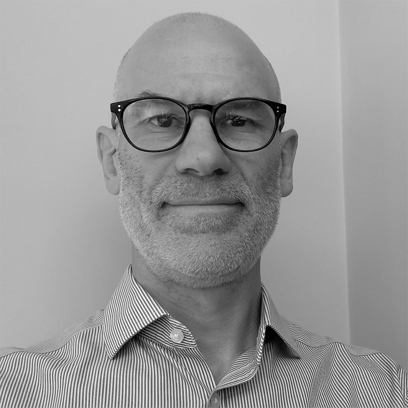 A bald man with glasses and a grey beard, wearing a striped shirt, is standing in front of a plain background.