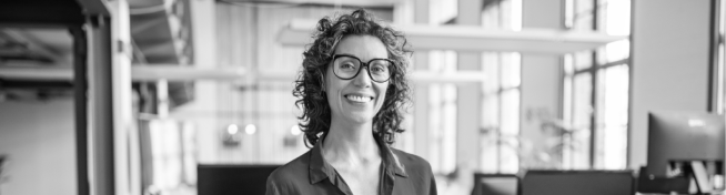 A person with curly hair and glasses smiles while standing in an office setting.