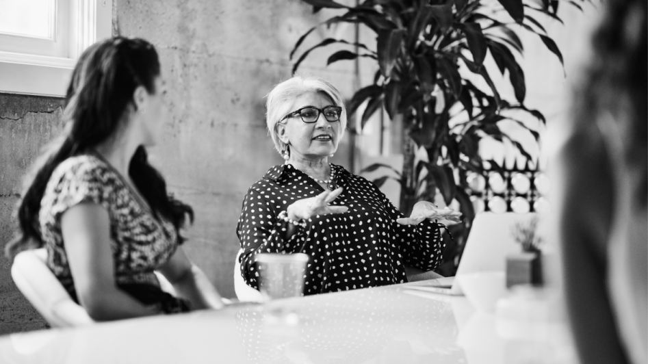 A woman in glasses speaks, gesturing with her hand, while sitting at a table with another woman in a patterned top. A laptop and a plant are in the background.