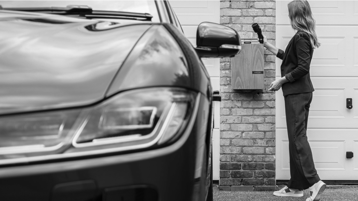 A woman in casual attire places mail in a brick wall mailbox next to a garage, with a car parked nearby. The image is in black and white.