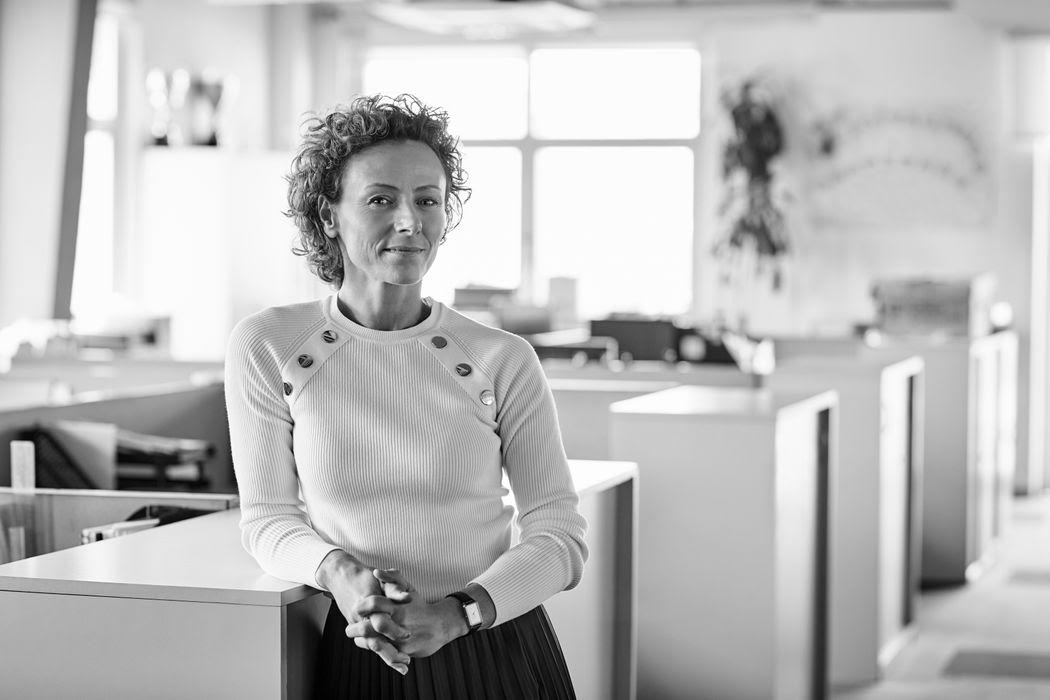 Person with curly hair stands leaning on a desk in an office environment, wearing a long-sleeve top.