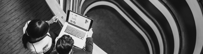 Two people viewing a laptop displaying charts, on a balcony with a downward view of winding staircases.