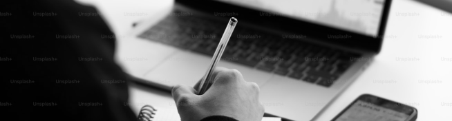 Person holding a pen near an open laptop and a smartphone on a desk.