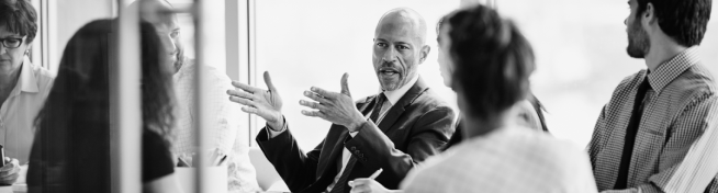 A group of people in a meeting room, with one man gesturing while speaking.