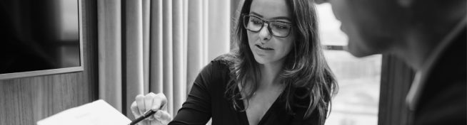 A woman wearing glasses discusses a document with a man in an office setting.