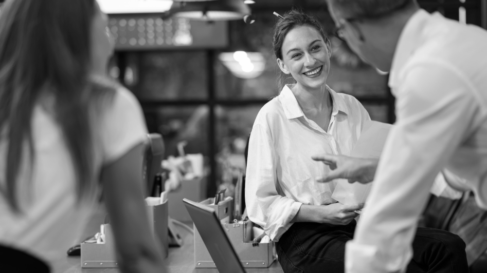 Three people in a casual office setting converse, with one woman smiling as a man gestures while speaking.