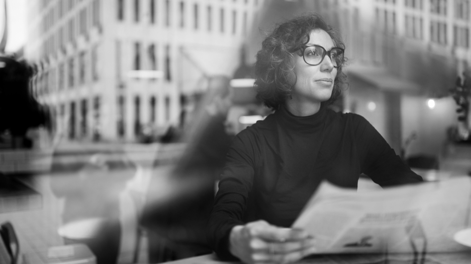 A woman with curly hair and glasses reads a newspaper at a table in a city setting, visible through a glass window.