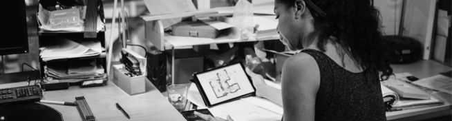 A woman works at a cluttered desk with papers and a tablet displaying a floor plan.