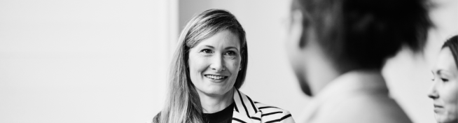 Three people engaged in a conversation; the focus is on a smiling woman wearing a striped blazer. The photo is in black and white.