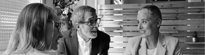 Three people are sitting and talking, with one person in glasses speaking animatedly. They are indoors, next to a wooden divider and a plant.