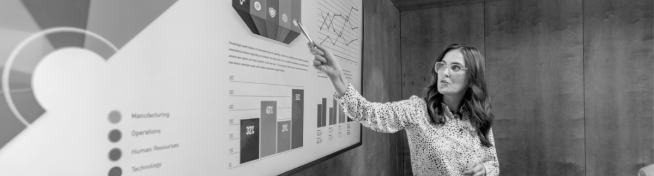 A woman wearing glasses points to a section of a pie chart on a large presentation screen in a meeting room.