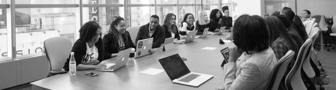 A group of people sitting around a large conference table with laptops and notepads, engaged in discussion in a modern office setting.