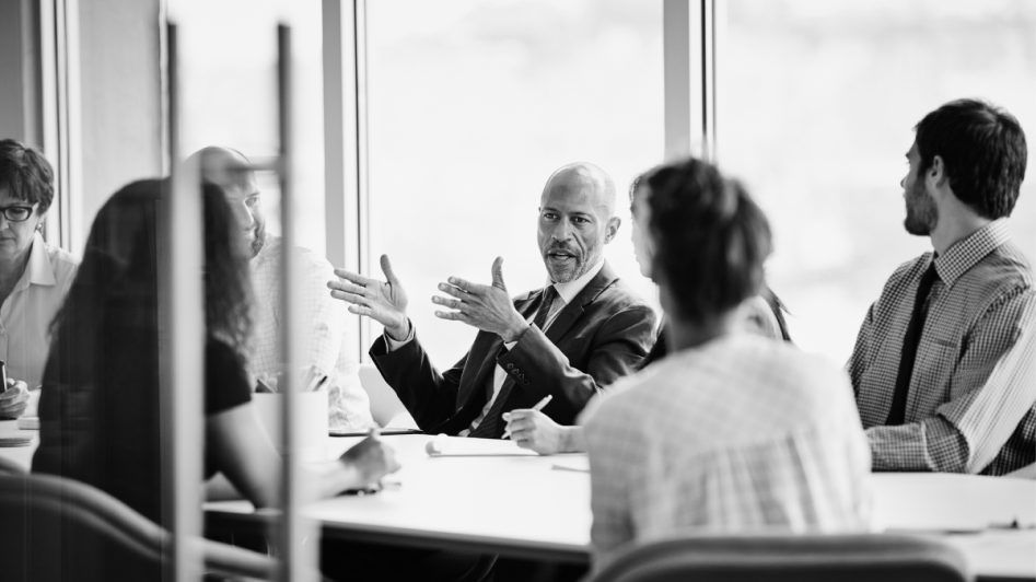 A group of people sits around a conference table in discussion, with a man in a suit gesturing as he speaks. The room has large windows showing an outdoor view.