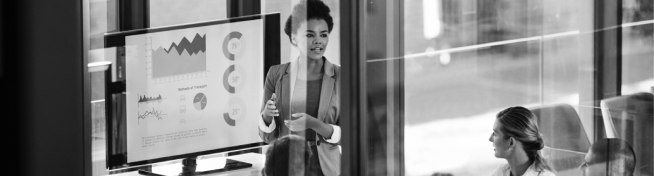 A person giving a presentation with graphs on a screen while two people watch attentively in a modern office setting.