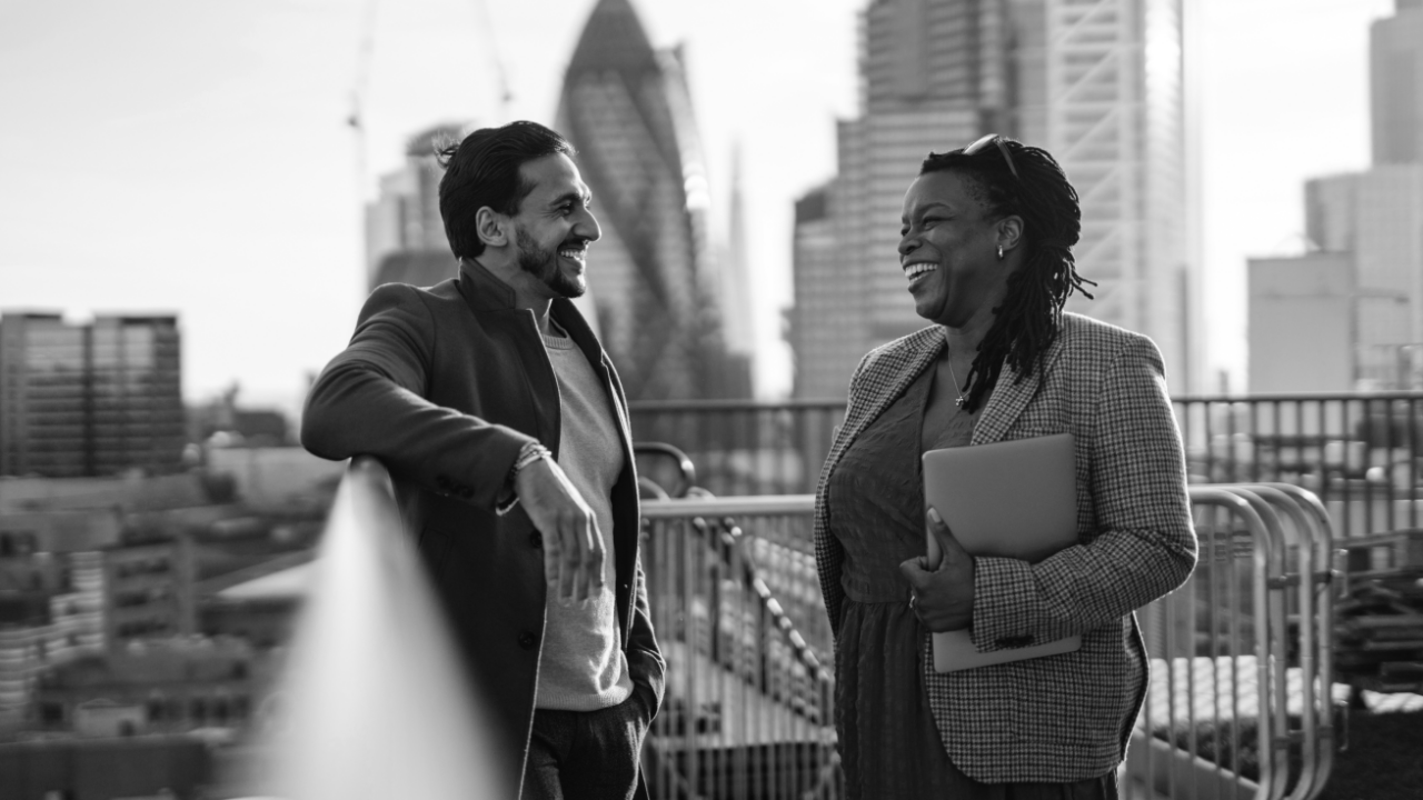 Two people stand on a rooftop, smiling and talking. City buildings are visible in the background. One holds a laptop.