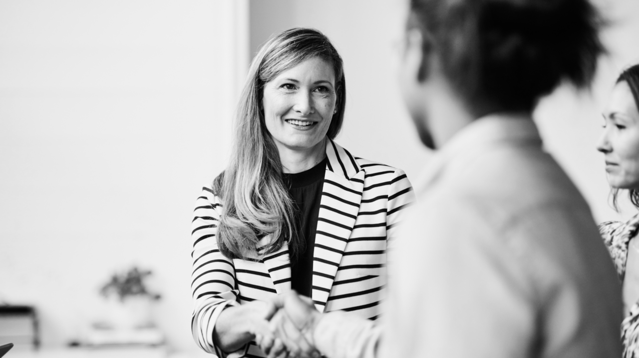 A person in a striped blazer smiles while shaking hands with another person in a meeting setting.