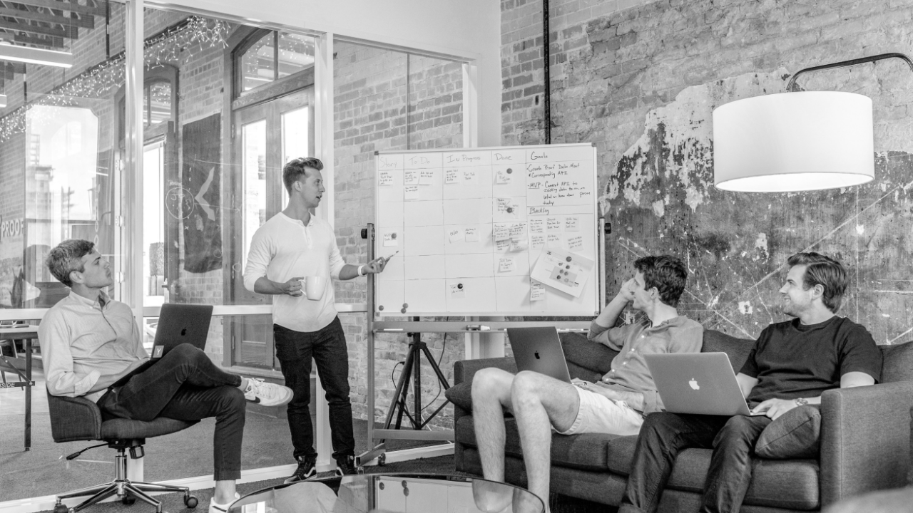 A group of four people in a meeting room, with one person standing and presenting information on a whiteboard. Three others are seated, using laptops and listening attentively.