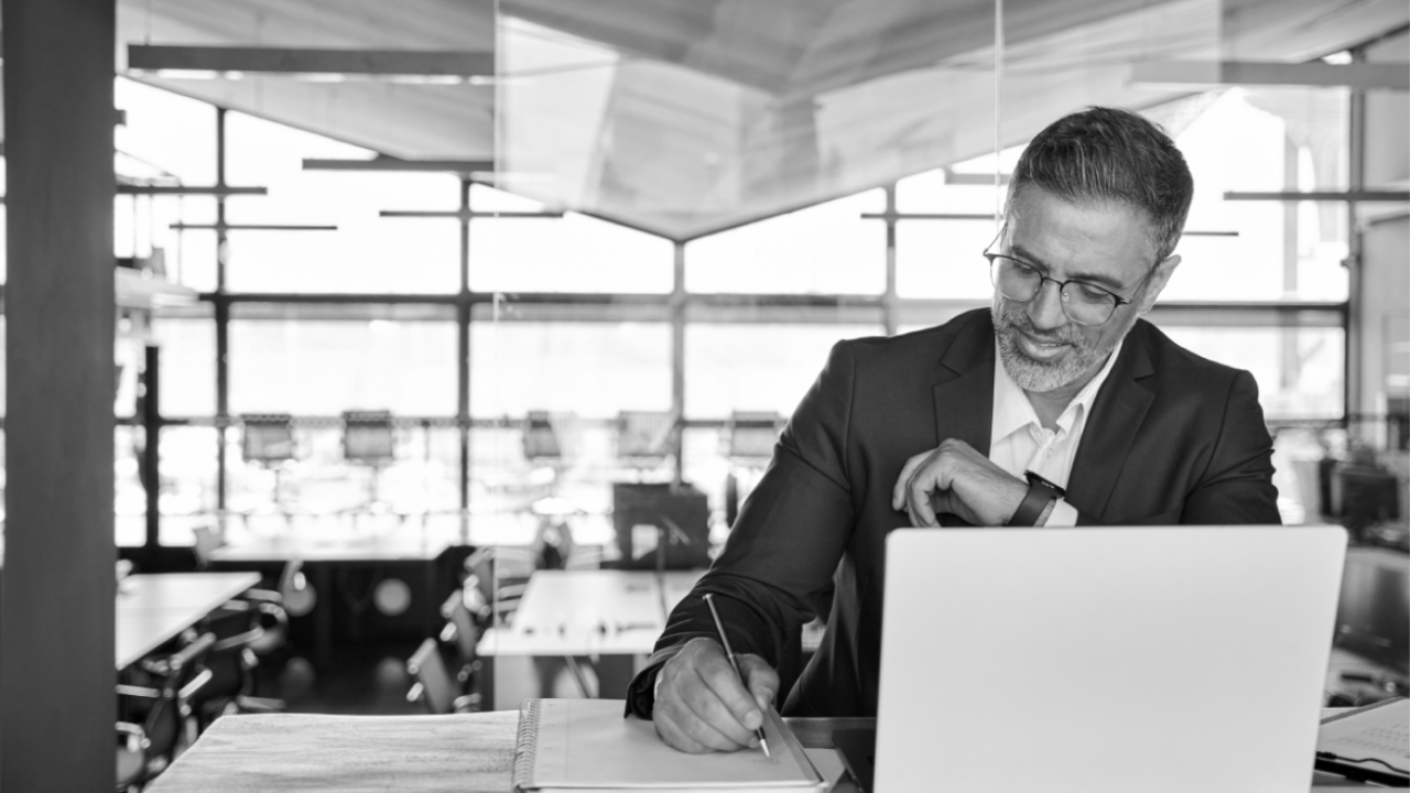 A man in a suit is seated at a desk, writing with a pen in a notebook while looking at a laptop. The background shows an office setting with large windows.