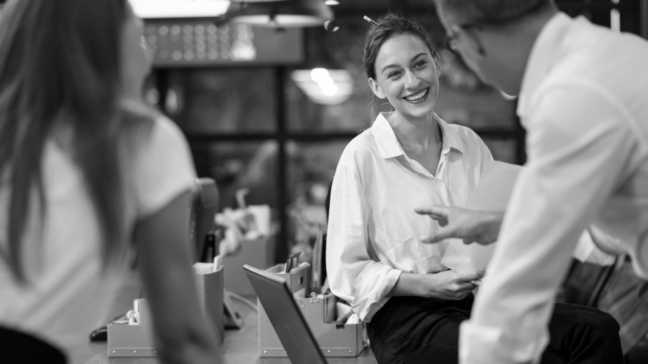 Three people engage in a conversation in a modern office, with one person sitting on a desk and smiling. The atmosphere appears casual and friendly.