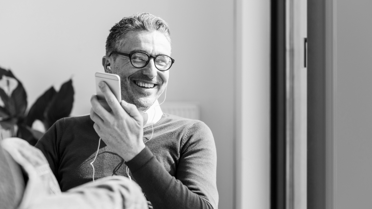 A man wearing glasses and earphones smiles while holding a smartphone.