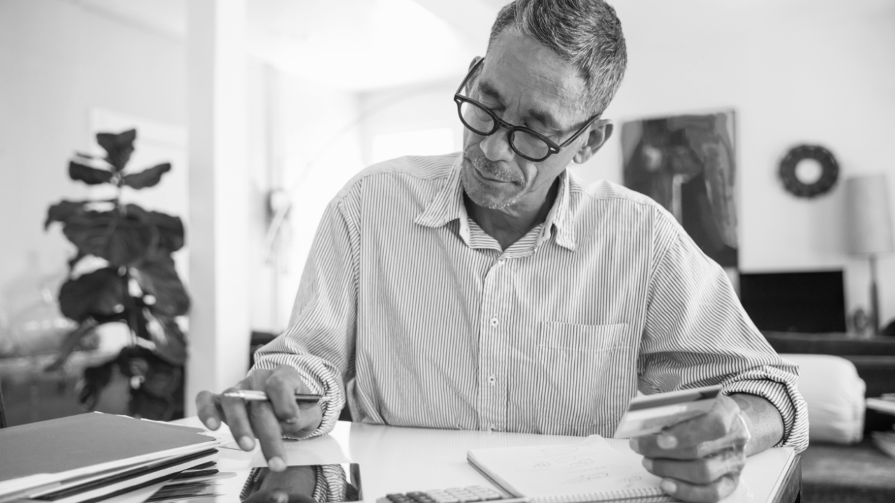 A man in glasses sits at a desk, writing in a notebook and holding a credit card, with papers and a calculator nearby.