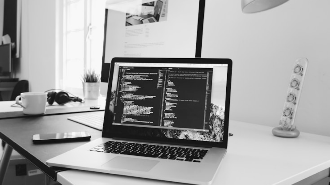 A laptop displaying code is on a desk with a smartphone, headphones, a cup, and a lamp.