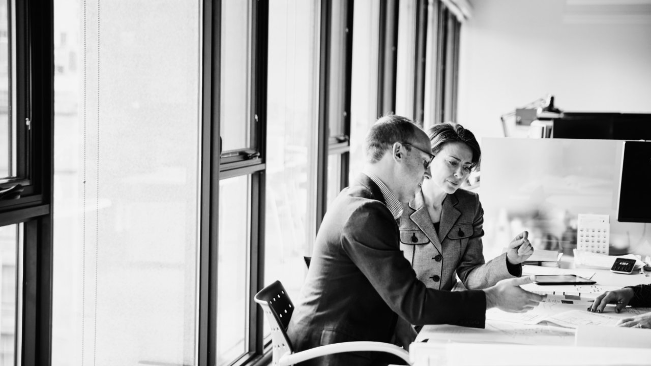 Two people in an office setting work together at a desk, examining documents and using a computer.