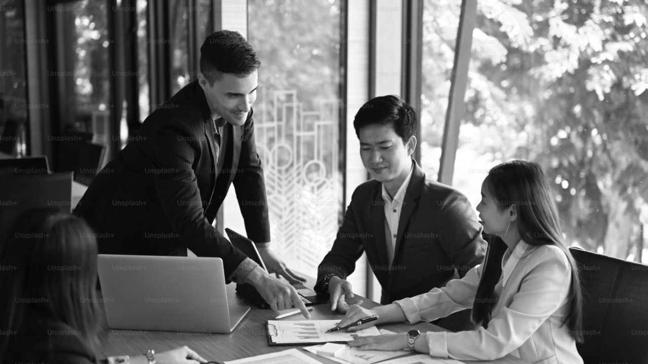 Four people are in a business meeting, seated around a table with papers and a laptop. One person is standing, pointing at a document.