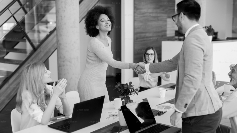 A woman and a man shake hands in an office meeting room while two other women sit at a table with laptops and papers.