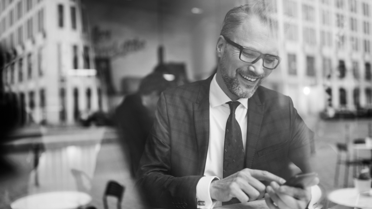 Black and white image of a man in a suit and glasses sitting at a table, smiling while using a smartphone. Reflections of buildings are visible in the background.