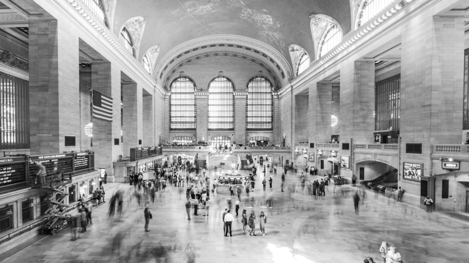 Black and white image of a busy train station interior with arched windows, high ceilings, and people walking on a large concourse. An American flag hangs on the left.