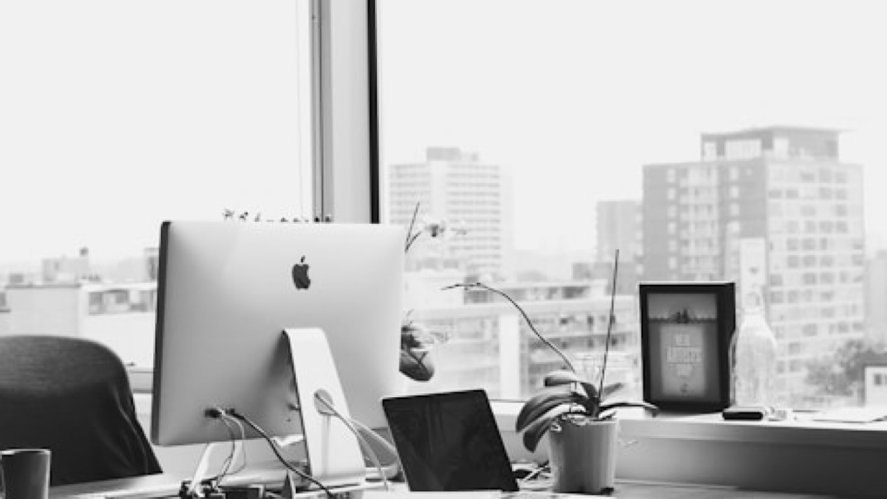 A grayscale image of an office desk with an iMac, a MacBook, a cup, a small plant, and a large window showing city buildings in the background.