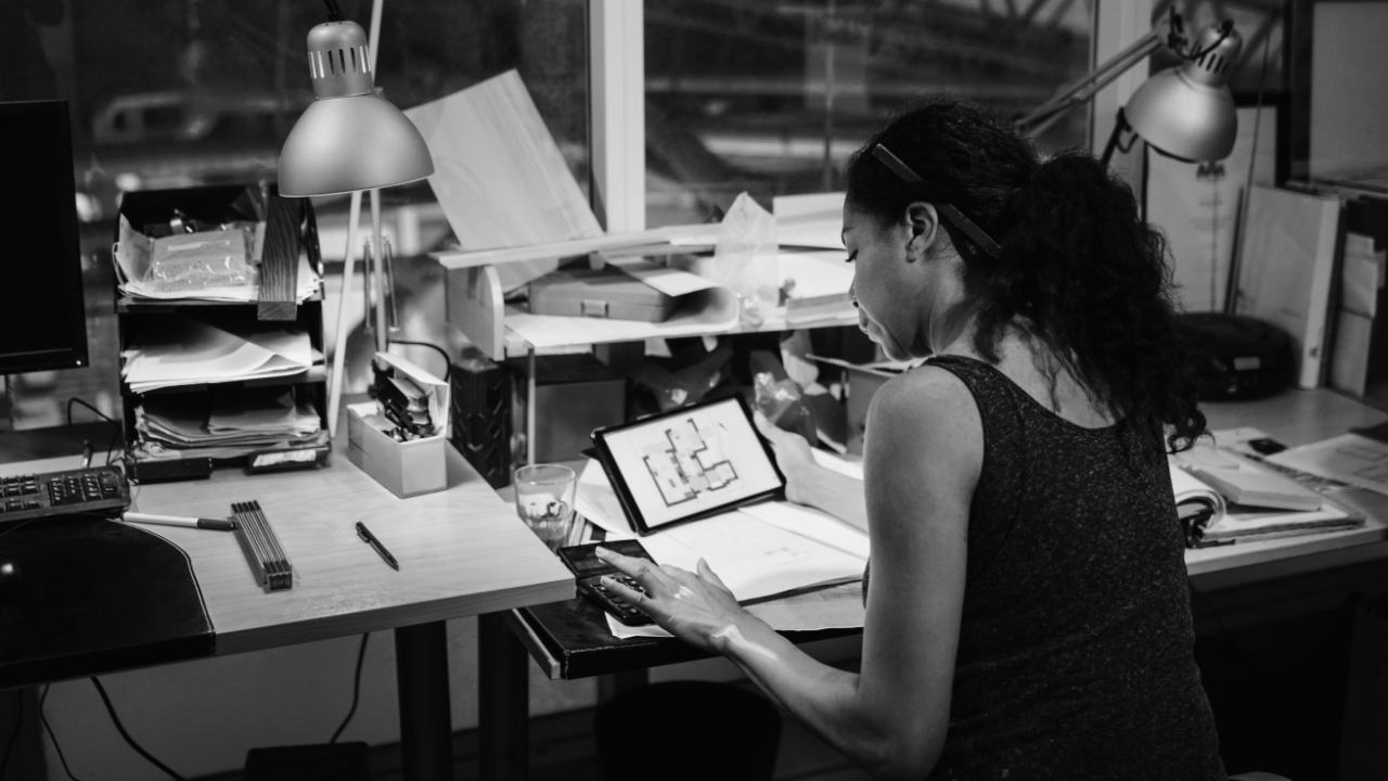 A person works at a desk with a tablet displaying a floor plan. The workspace is cluttered with papers, books, and two desk lamps.