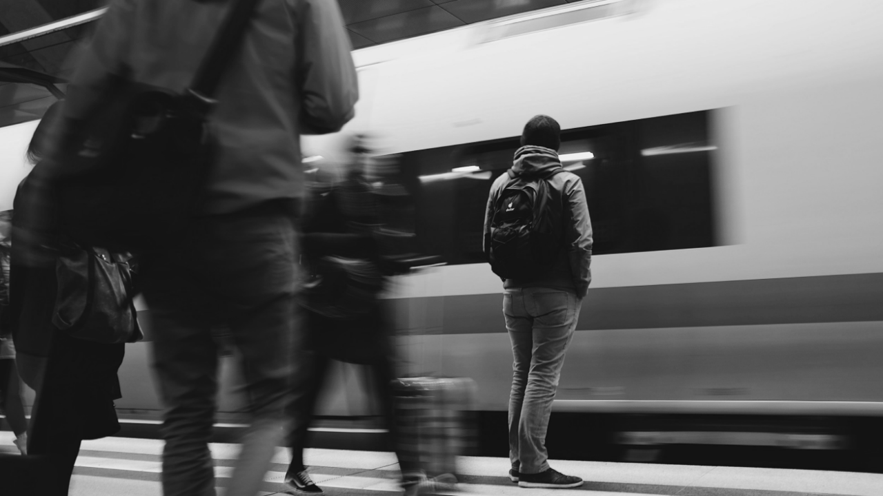 Person standing on a train platform with a backpack, watching a passing train in motion. The scene is in black and white.