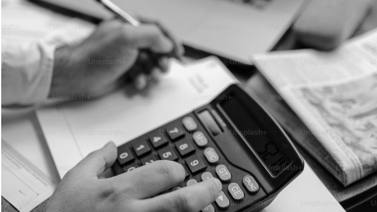 A person uses a calculator while taking notes on a document, with a newspaper partially visible on the desk.