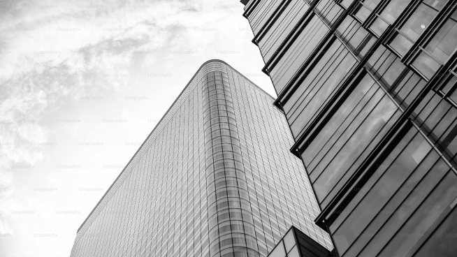 Black and white photo of a modern building with glass windows and reflective surfaces, set against a cloudy sky.