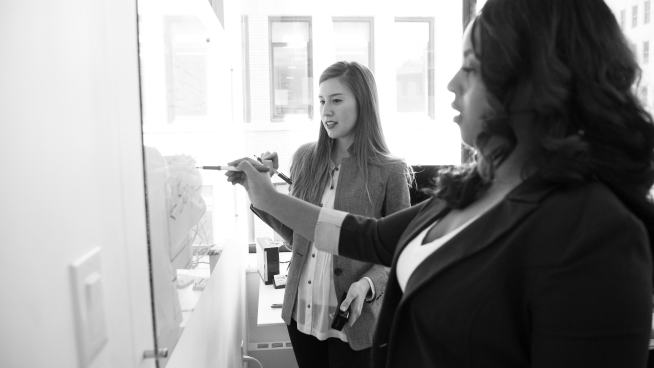 Two women stand in an office, discussing and writing notes on a glass wall.