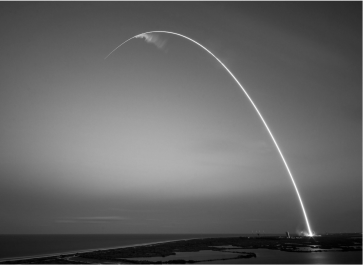 A long-exposure photograph showing a rocket's bright arc during its ascent over a coastal landscape at dusk.