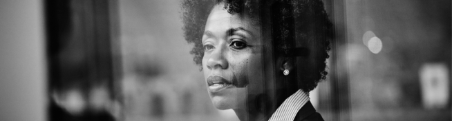 Black and white image of a person with curly hair and a thoughtful expression, viewed through a glass panel.