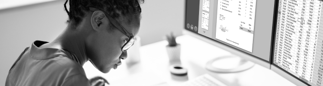 Person wearing glasses focused on computer screens displaying spreadsheets and documents.