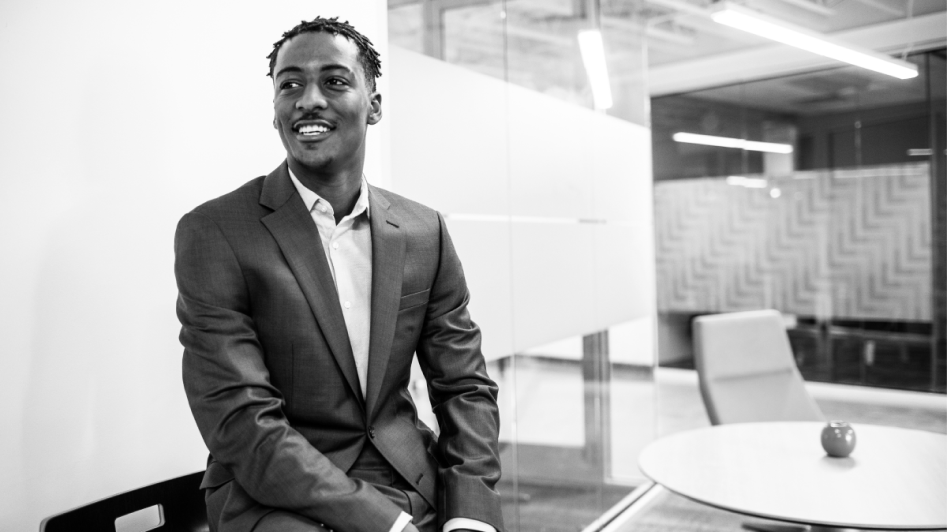 A man in a suit sits on a chair in a modern office, smiling.