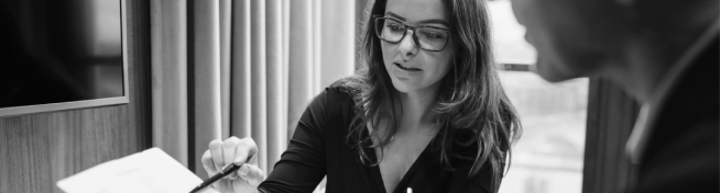 A woman wearing glasses is sitting at a table and pointing at a document. She appears to be discussing something with another person whose face is not fully visible.