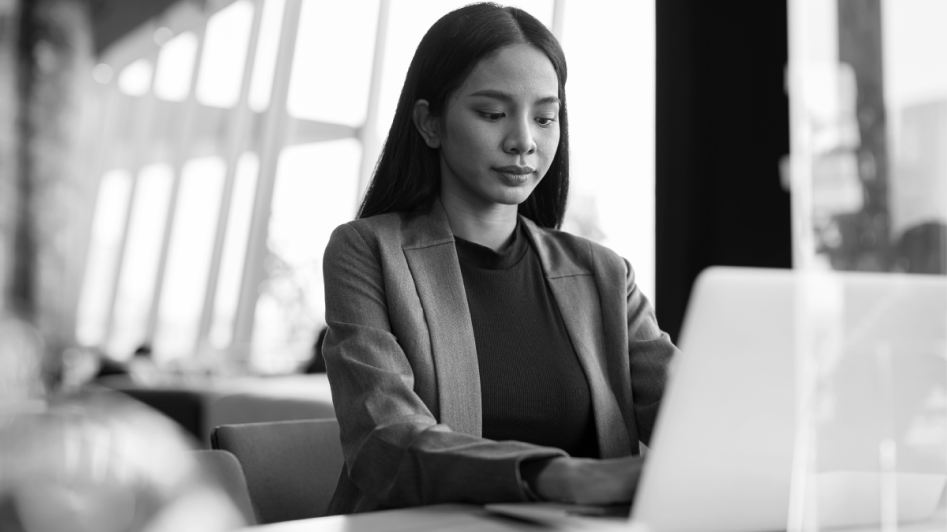 A woman in a blazer works on a laptop at a desk in a bright, modern office.