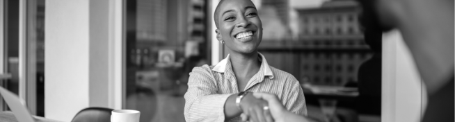 A person smiles while shaking hands in an office setting with a cup and a laptop on the table.