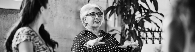 A woman with glasses and a polka dot blouse gestures while speaking to another woman in a patterned shirt. They are in a room with a concrete wall and plants.