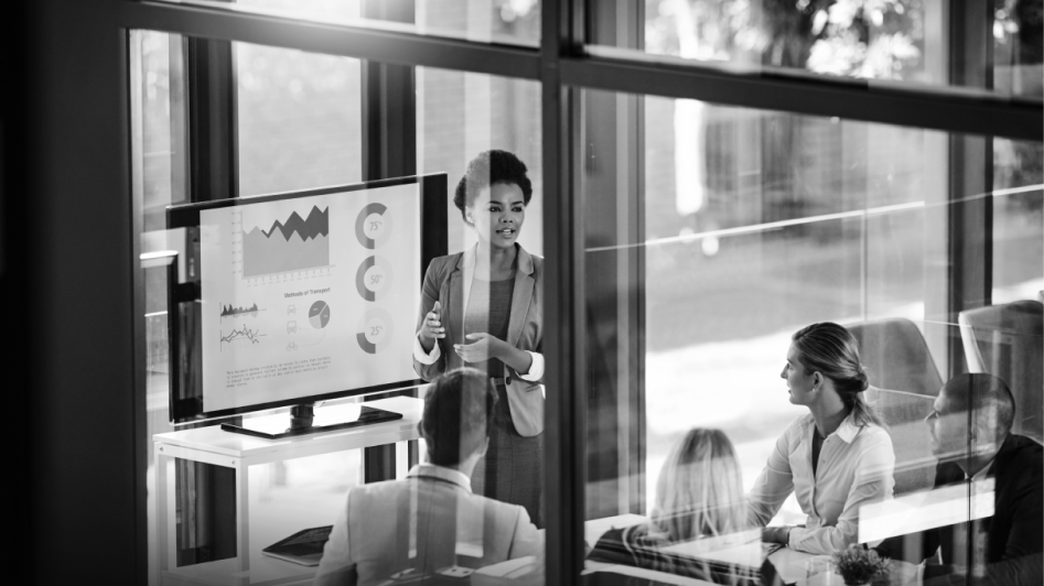 A person presents data on a screen to a group in a glass-walled meeting room.