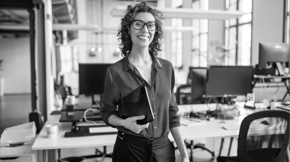 A person with curly hair and glasses stands smiling in a modern office, holding a tablet.