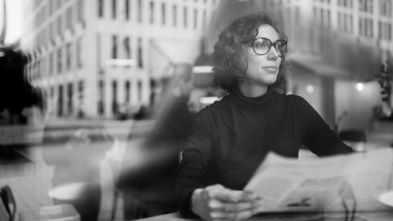 A woman with glasses sits at a table by a window, holding a newspaper and gazing outside. The reflection shows a cityscape with tall buildings. Black and white photo.