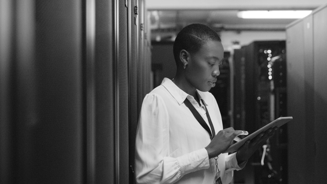 A person in a server room, wearing a white shirt, is using a tablet.