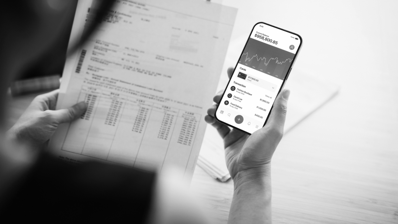 A person holding a smartphone displaying stock market data in one hand and reviewing financial documents in the other.